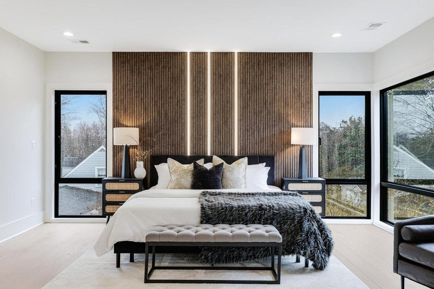 Modern bedroom design featuring a king bed with layered pillows, tufted bench, black nightstands with lamps, and a wood slat accent wall with built-in LED lighting between large black-frame windows.