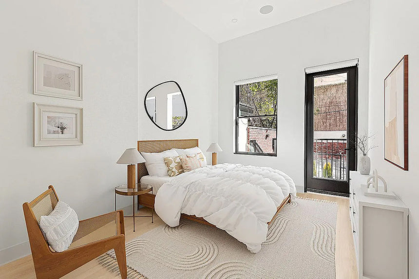 Bright modern bedroom with cane bed frame, white bedding, wood accent chair, and large window for natural light.