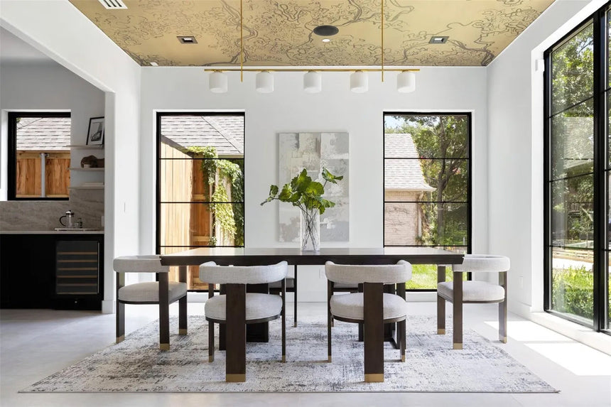 Elegant dining room with patterned gold ceiling, large black-framed windows, and a rectangular wood dining table.