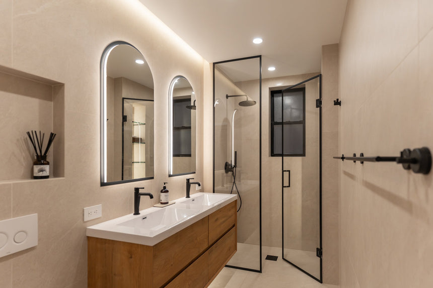 Modern bathroom with a floating wood double-sink vanity, matte black faucets, LED arched mirrors, and a frameless glass shower.