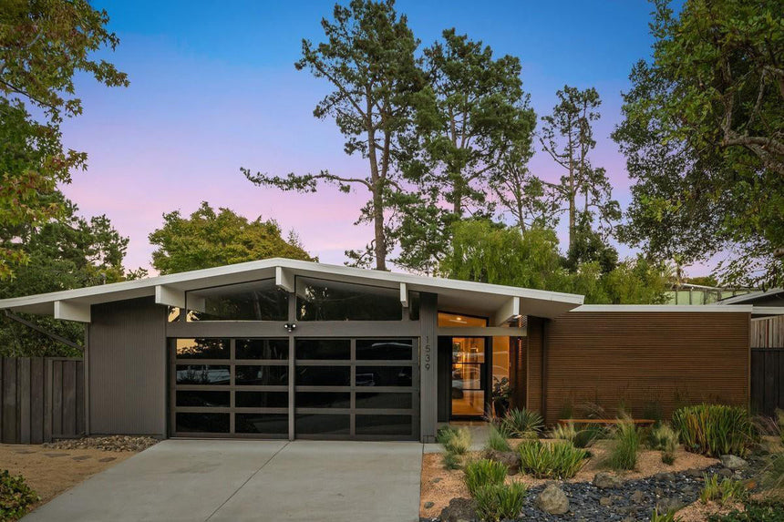 Modern mid-century home exterior with large glass garage doors, sloped roofline, and landscaped front yard at sunset.