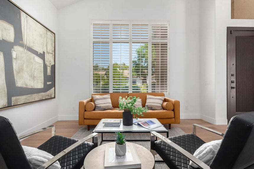 Modern Scottsdale living room with tall windows, white walls, and a camel-tan leather sofa accented by neutral pillows and contemporary abstract art.