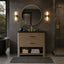 A modern bathroom vanity with a woven natural-fiber cabinet, black countertop, dual brass faucets, a round gold-framed mirror, and matching wall sconces, shown in a dark stone bathroom with a glass shower enclosure.