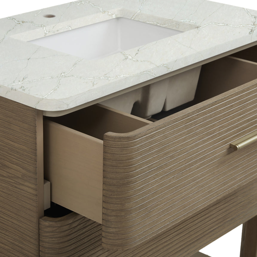 A modern bathroom vanity with a light oak wood frame, six drawers, an open lower shelf, and a white marble top with an integrated sink. The vanity is styled in a bright bathroom with a white freestanding tub, a gold faucet, and a glass shower stall in the background.