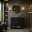 A modern bathroom vanity setup with a black textured double-drawer cabinet, gold handles, a matching gold faucet, a round gold-framed mirror, and warm wall sconces in a dark stone bathroom.