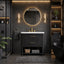 A modern bathroom vanity setup with a black textured cabinet, white sink countertop, gold faucet and handles, a round backlit mirror, and warm wall sconces in a dark charcoal bathroom.