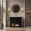 A modern bathroom vanity setup featuring a black textured cabinet with gold handles, a white sink basin, a gold faucet, a round gold-framed mirror, and matching wall sconces in a warm neutral bathroom setting.