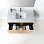 Top-down view of a modern bathroom vanity with a light gray countertop, integrated rectangular sink, black faucet, and open wood-grain drawer containing toiletries, set against a white wall and floor.