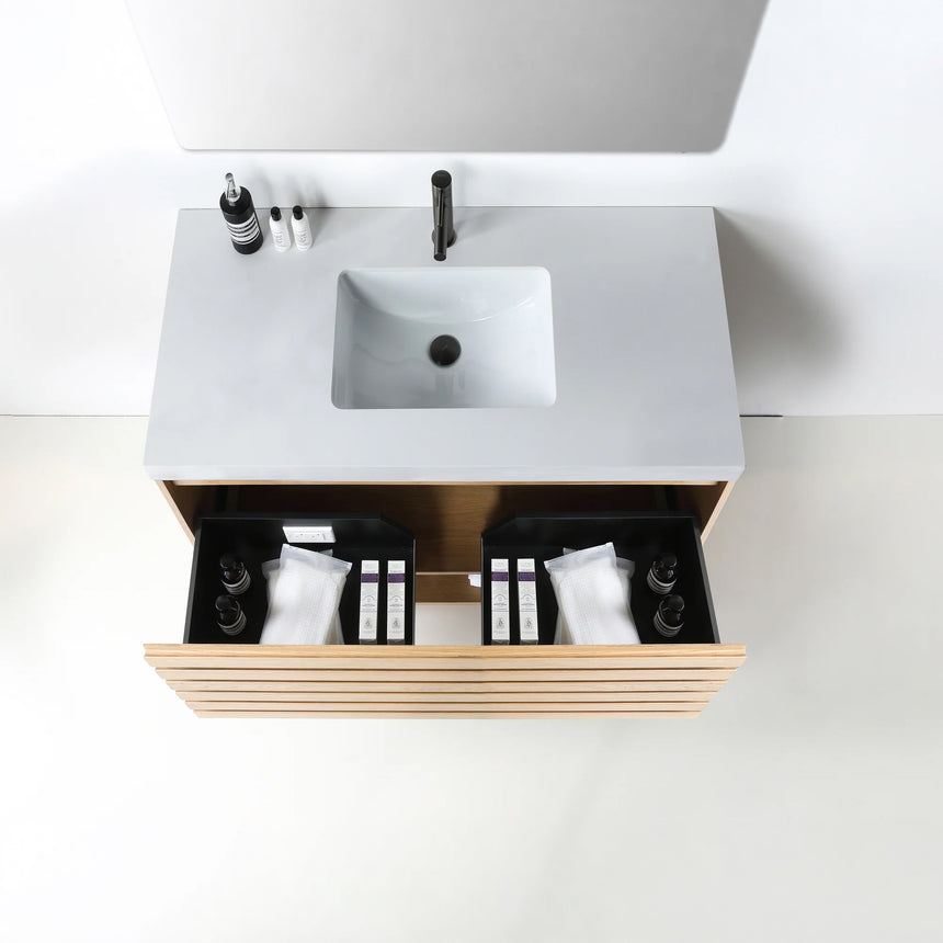 A modern floating bathroom vanity with a light oak wood finish, a white countertop, and an integrated sink, shown in a styled bathroom setting with a bathtub, shower glass, and decorative plants.