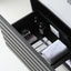 An open dark gray bathroom drawer showing toiletries including a white folded towel, boxed items, and two dark bottles, photographed on a clean white background.