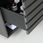 A close-up of a dark gray wooden drawer with a slatted front, shown partially open to reveal a black interior shelf holding small bottles and a folded white cloth.