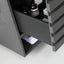 A close-up of a dark gray wooden drawer unit with one drawer pulled open, showing interior storage space with amber bottles and a white box. The drawer front has a distinctive wavy, grooved texture.