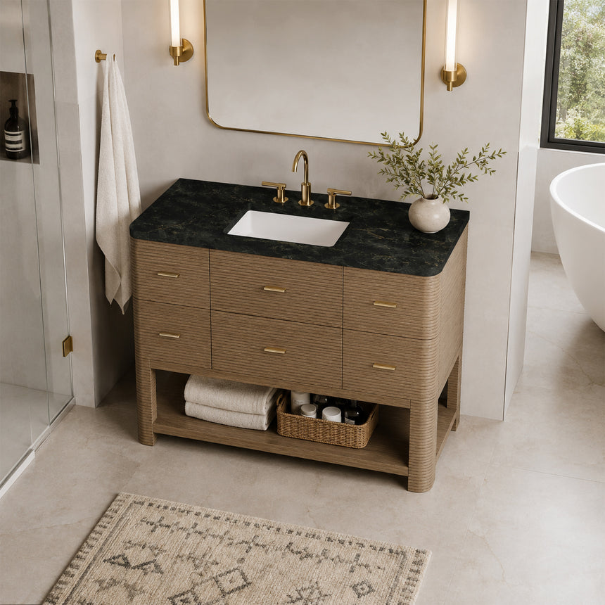 A modern bathroom vanity with a light oak wood frame, six drawers, an open lower shelf, and a white marble top with an integrated sink. The vanity is styled in a bright bathroom with a white freestanding tub, a gold faucet, and a glass shower stall in the background.