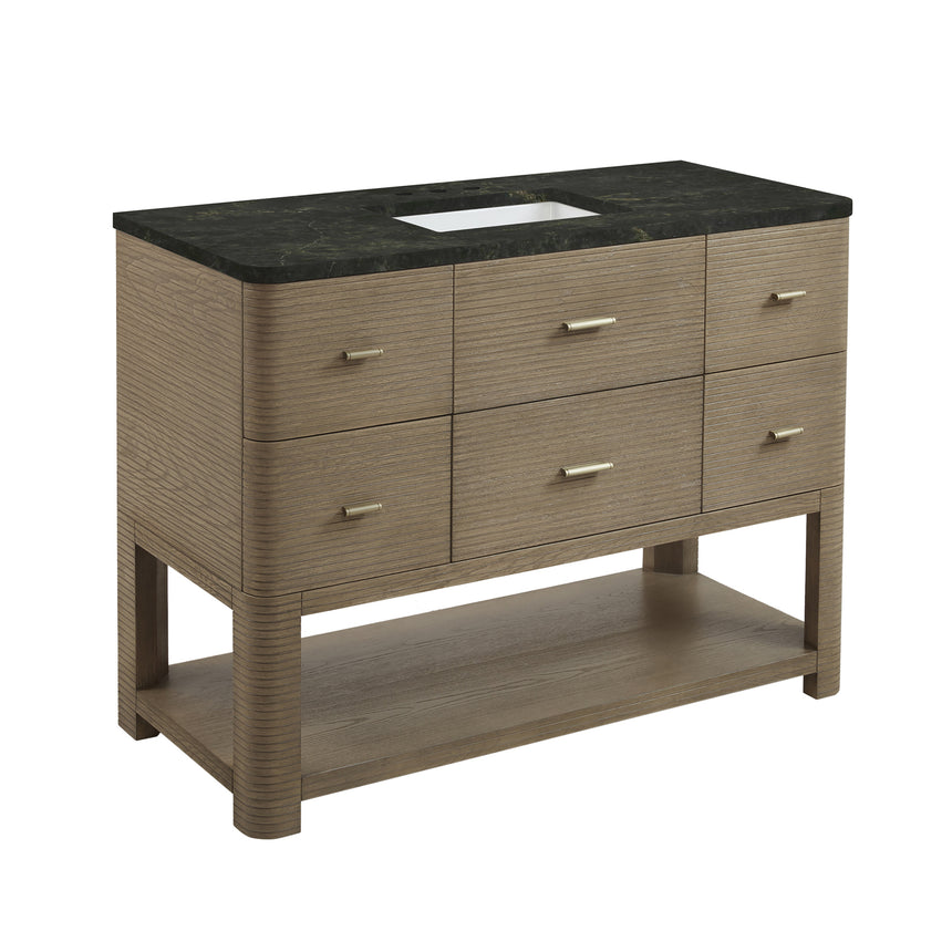 A modern bathroom vanity with a light oak wood frame, six drawers, an open lower shelf, and a white marble top with an integrated sink. The vanity is styled in a bright bathroom with a white freestanding tub, a gold faucet, and a glass shower stall in the background.