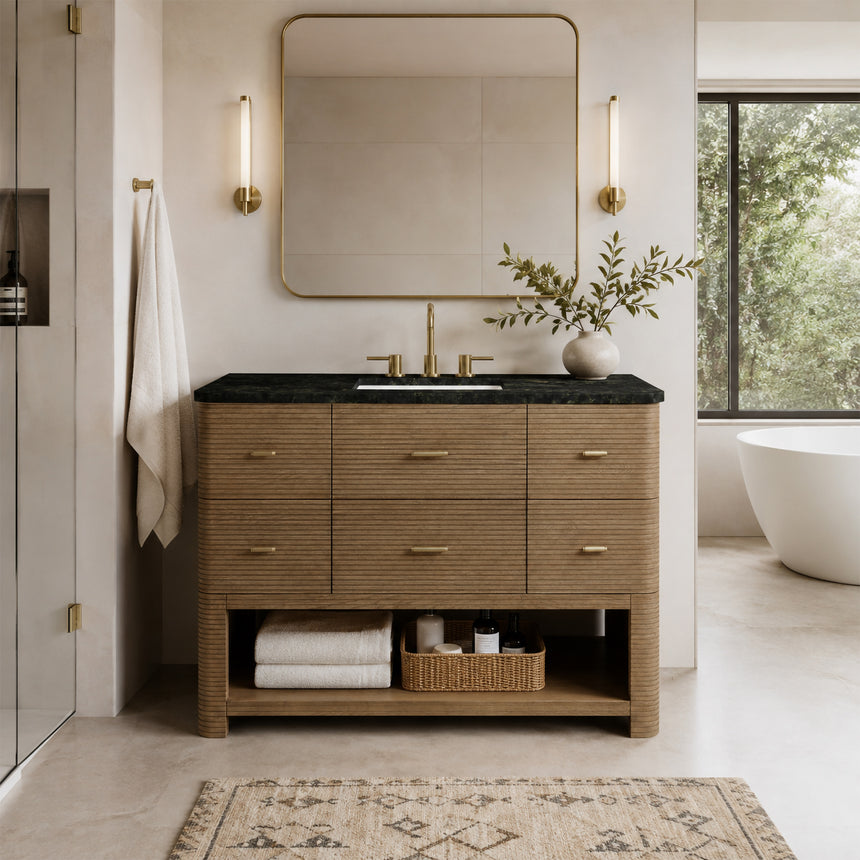 A modern bathroom vanity with a light oak wood frame, six drawers, an open lower shelf, and a white marble top with an integrated sink. The vanity is styled in a bright bathroom with a white freestanding tub, a gold faucet, and a glass shower stall in the background.