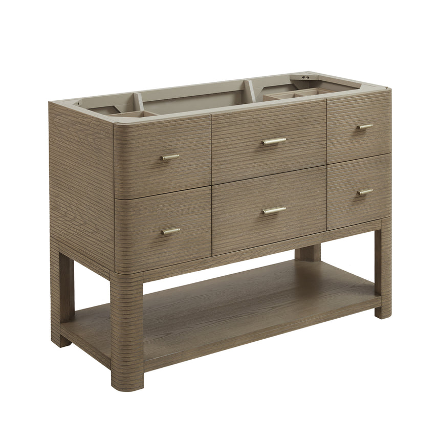 A modern bathroom vanity with a light oak wood frame, six drawers, an open lower shelf, and a white marble top with an integrated sink. The vanity is styled in a bright bathroom with a white freestanding tub, a gold faucet, and a glass shower stall in the background.