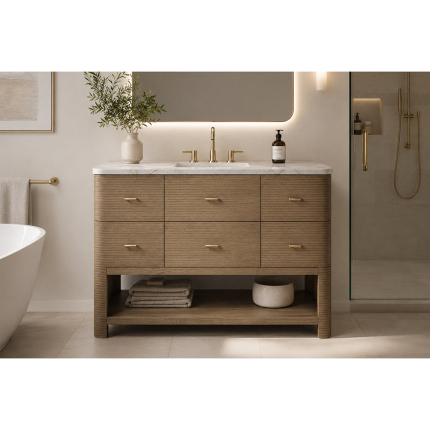 A modern bathroom vanity with a light oak wood frame, six drawers, an open lower shelf, and a white marble top with an integrated sink. The vanity is styled in a bright bathroom with a white freestanding tub, a gold faucet, and a glass shower stall in the background.