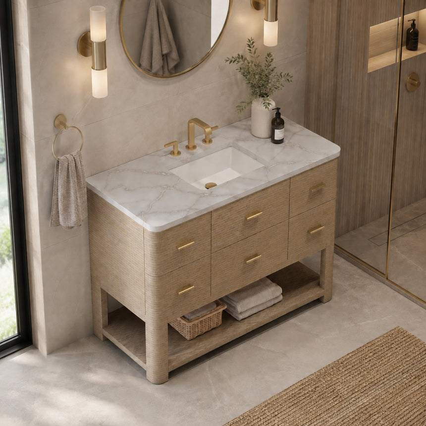 A modern bathroom vanity with a light oak wood frame, six drawers, an open lower shelf, and a white marble top with an integrated sink. The vanity is styled in a bright bathroom with a white freestanding tub, a gold faucet, and a glass shower stall in the background.