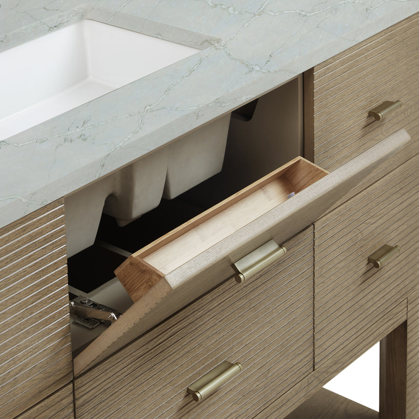 A modern bathroom vanity with a light oak wood frame, six drawers, an open lower shelf, and a white marble top with an integrated sink. The vanity is styled in a bright bathroom with a white freestanding tub, a gold faucet, and a glass shower stall in the background.