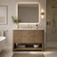 A modern bathroom vanity with a light wood textured cabinet, white marble countertop, and a large rectangular backlit mirror. The vanity is styled with a gold faucet, a vase of greenery, and a bottle of soap, set against a neutral beige wall with a glass shower enclosure on the right.