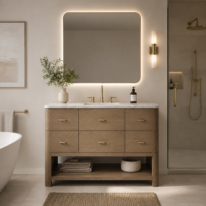 A modern bathroom vanity with a light oak wood frame, six drawers, an open lower shelf, and a white marble top with an integrated sink. The vanity is styled in a bright bathroom with a white freestanding tub, a gold faucet, and a glass shower stall in the background.