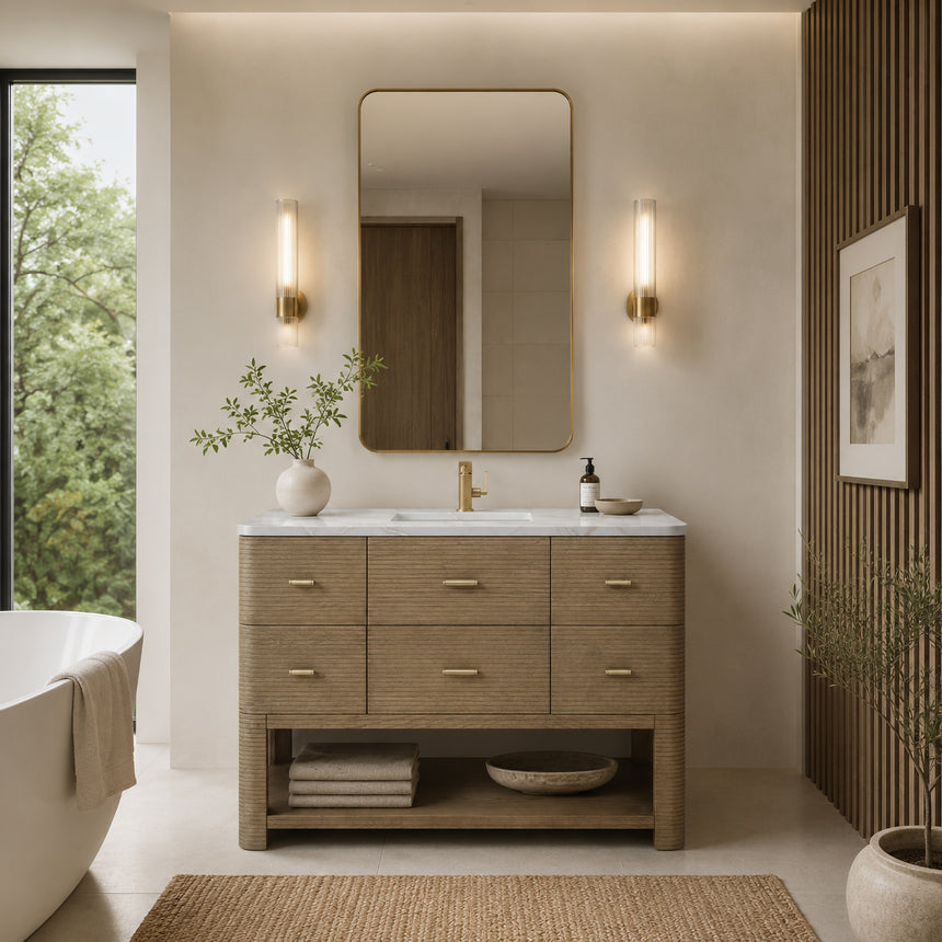 A modern bathroom vanity with a light oak wood frame, six drawers, an open lower shelf, and a white marble top with an integrated sink. The vanity is styled in a bright bathroom with a white freestanding tub, a gold faucet, and a glass shower stall in the background.