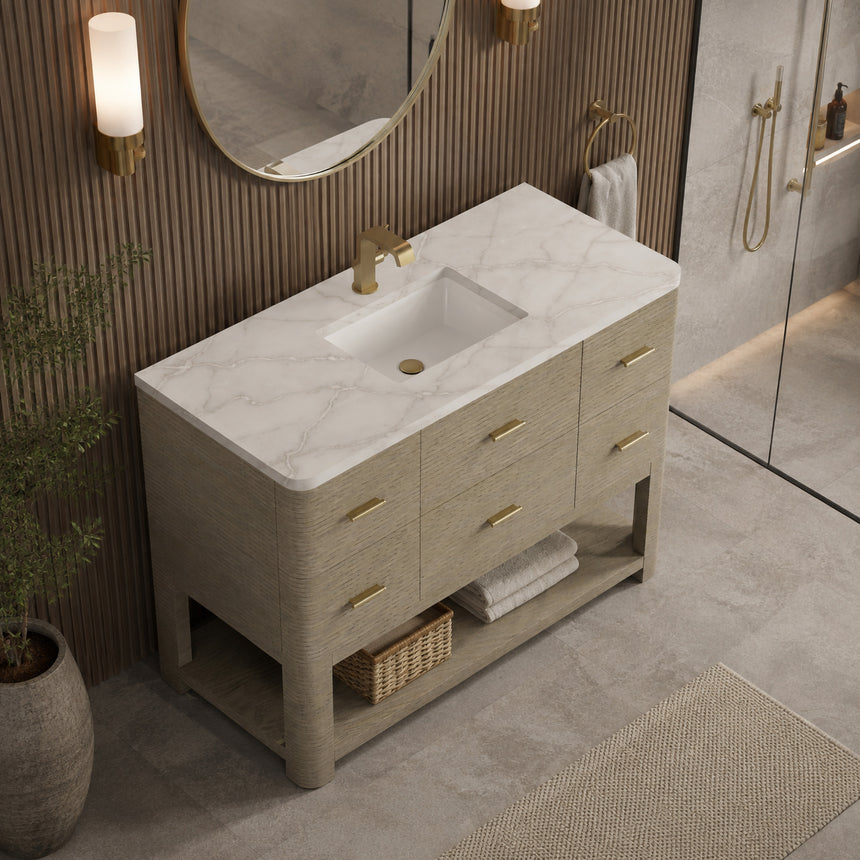 A modern bathroom vanity with a light oak wood frame, six drawers, an open lower shelf, and a white marble top with an integrated sink. The vanity is styled in a bright bathroom with a white freestanding tub, a gold faucet, and a glass shower stall in the background.