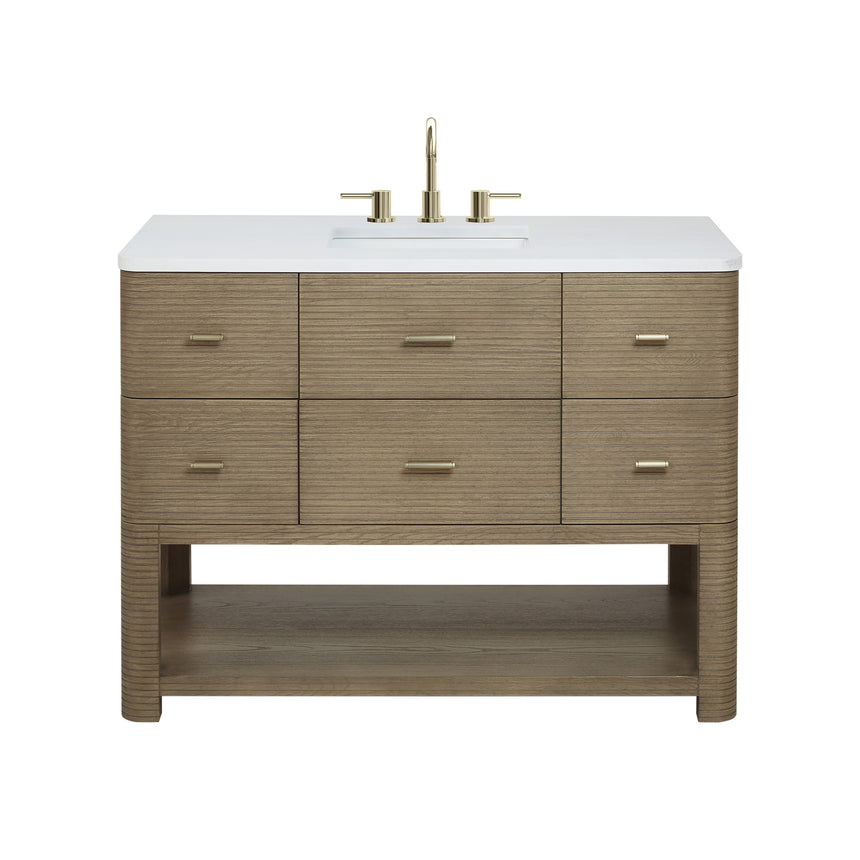 A modern bathroom vanity with a light oak wood frame, six drawers, an open lower shelf, and a white marble top with an integrated sink. The vanity is styled in a bright bathroom with a white freestanding tub, a gold faucet, and a glass shower stall in the background.