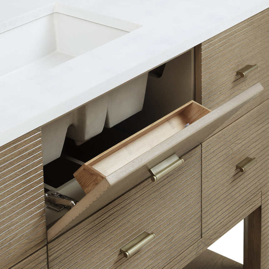A modern bathroom vanity with a light oak wood frame, six drawers, an open lower shelf, and a white marble top with an integrated sink. The vanity is styled in a bright bathroom with a white freestanding tub, a gold faucet, and a glass shower stall in the background.