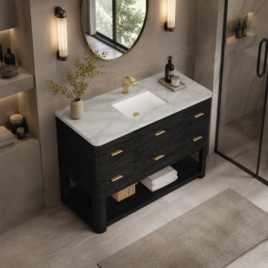 A modern bathroom vanity with a light oak wood frame, six drawers, an open lower shelf, and a white marble top with an integrated sink. The vanity is styled in a bright bathroom with a white freestanding tub, a gold faucet, and a glass shower stall in the background.