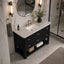 A modern bathroom vanity with a black wood cabinet, white marble countertop, and gold hardware, shown in a styled bathroom setting with a round mirror, wall sconces, and a glass shower door.