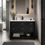 A modern bathroom vanity with a black textured cabinet, white countertop, matte black sink and faucet, and gold-tone drawer pulls, shown in a styled bathroom setting with a mirror, wall sconces, towels, and a rug.