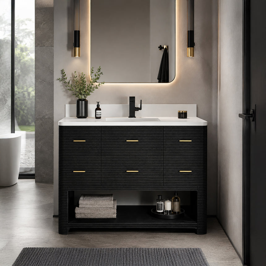 A modern bathroom vanity with a light oak wood frame, six drawers, an open lower shelf, and a white marble top with an integrated sink. The vanity is styled in a bright bathroom with a white freestanding tub, a gold faucet, and a glass shower stall in the background.