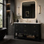 A modern bathroom vanity with a black ribbed cabinet, white countertop, gold faucet and handles, and a large backlit rectangular mirror. The scene is styled in a dark, contemporary bathroom with a black freestanding tub edge, built-in shelving, and wall sconces.