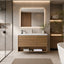 A modern bathroom vanity with a light wood floating cabinet, white countertop, and an integrated rectangular mirror with warm LED lighting. The bathroom features a glass shower enclosure on the left, a woven rug on the floor, and a white freestanding bathtub on the right.