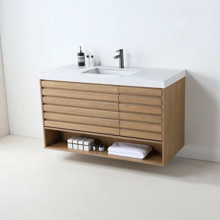 A modern floating bathroom vanity with a light oak wood finish, a white countertop, and an integrated sink, shown in a styled bathroom setting with a bathtub, shower glass, and decorative plants.