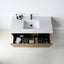 A modern wall-mounted bathroom vanity with a white rectangular sink, matte black faucet, and open wood-grain lower cabinet drawer showing toiletries, photographed from above against a clean white wall and floor.