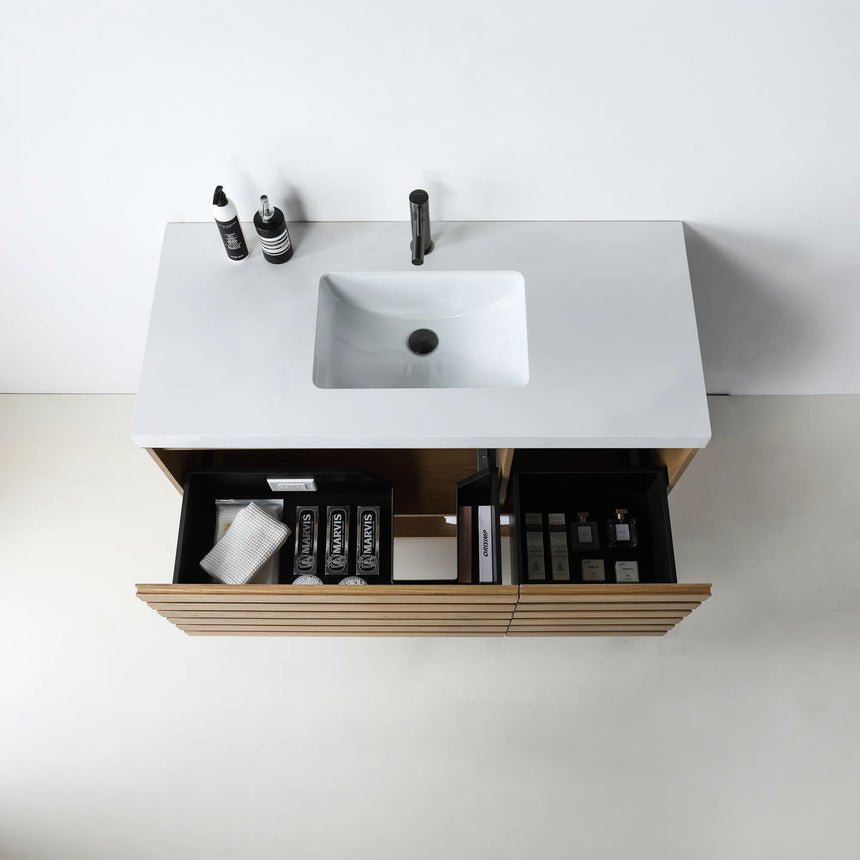 A modern floating bathroom vanity with a light oak wood finish, a white countertop, and an integrated sink, shown in a styled bathroom setting with a bathtub, shower glass, and decorative plants.