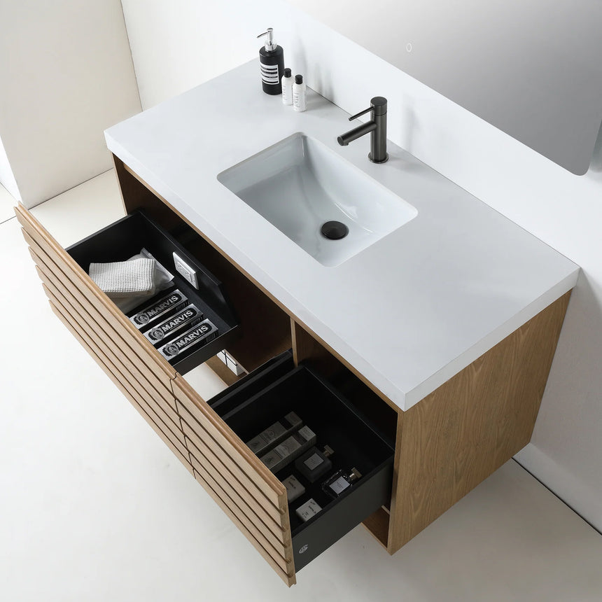 A modern floating bathroom vanity with a light oak wood finish, a white countertop, and an integrated sink, shown in a styled bathroom setting with a bathtub, shower glass, and decorative plants.