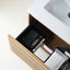 A close-up of an open wooden vanity drawer containing black and white storage organizers with visible MARVIS branding, next to a white sink countertop.