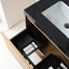 An open bathroom vanity drawer with a black organizer tray holding toiletries, shown next to a black marble countertop and white sink in a modern bathroom setting.