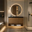 A modern bathroom vanity setup with a rectangular wood-look floating cabinet, black countertop, black sink, and a large round backlit mirror. A glass shower enclosure is visible on the right, and warm under-cabinet lighting illuminates the floor and shelf below.