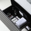 An open dark drawer in a bathroom or vanity setting, containing toiletries, a folded white towel, and a white outlet panel on the inside of the drawer.