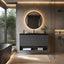 A modern bathroom vanity setup with a dark floating cabinet, black countertop, round backlit mirror, and open shelf storage, shown in a styled interior with a glass shower screen and warm ambient lighting.