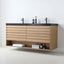 A modern wall-mounted bathroom vanity with a light oak wood slat front, open lower shelf, and a dark stone countertop holding two black faucets and small toiletries, shown against a white wall and floor.