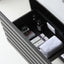 A dark charcoal gray bathroom drawer with a modern horizontal slat design, shown open to reveal toiletries including a white towel, bottles, and boxes inside.