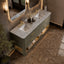 A modern double-sink bathroom vanity with a textured sage green cabinet, white marble countertops, and gold hardware, shown in a styled bathroom setting with marble floors and wall sconces.