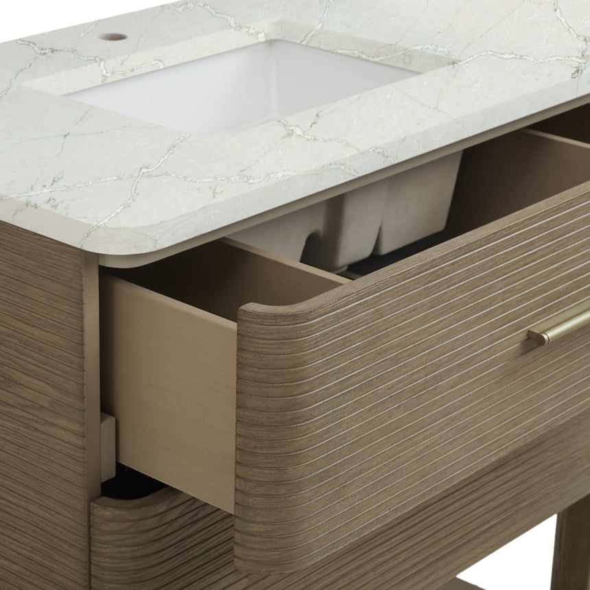 A modern bathroom vanity with a light oak wood frame, six drawers, an open lower shelf, and a white marble top with an integrated sink. The vanity is styled in a bright bathroom with a white freestanding tub, a gold faucet, and a glass shower stall in the background.
