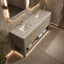 A modern double-sink bathroom vanity with a white marble-look countertop, textured beige cabinet, and gold handles, shown in a luxury bathroom setting with warm ambient lighting.