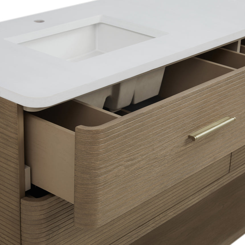 A modern bathroom vanity with a light oak wood frame, six drawers, an open lower shelf, and a white marble top with an integrated sink. The vanity is styled in a bright bathroom with a white freestanding tub, a gold faucet, and a glass shower stall in the background.